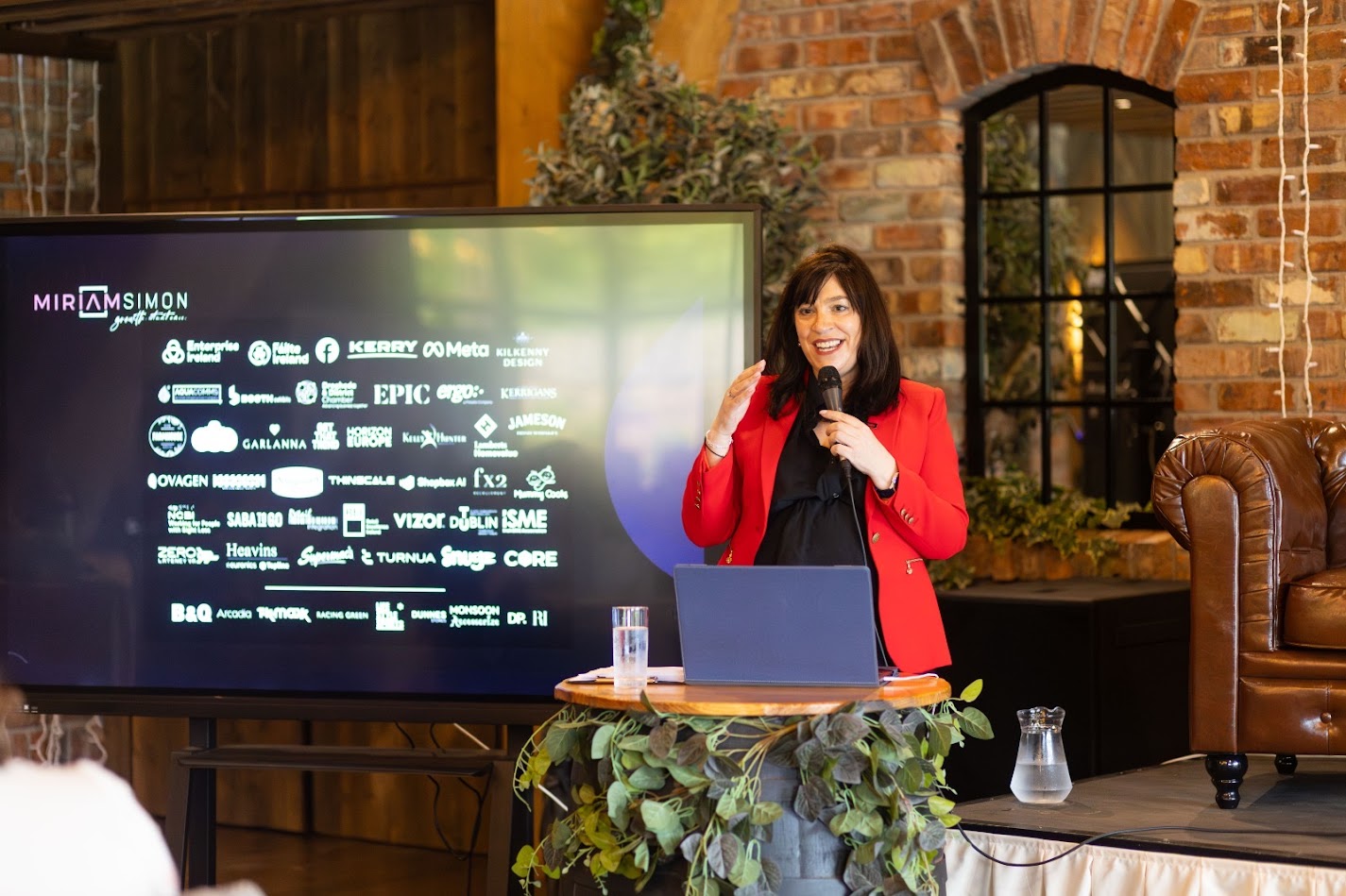Miriam Simon speaking on stage, microphone in hand, with her client logo wall on the screen behind her